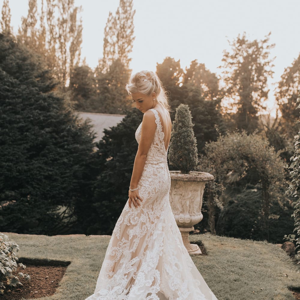 A bride in a long lace wedding dress stands in gardens for Crab & Lobster fairytale wedding.