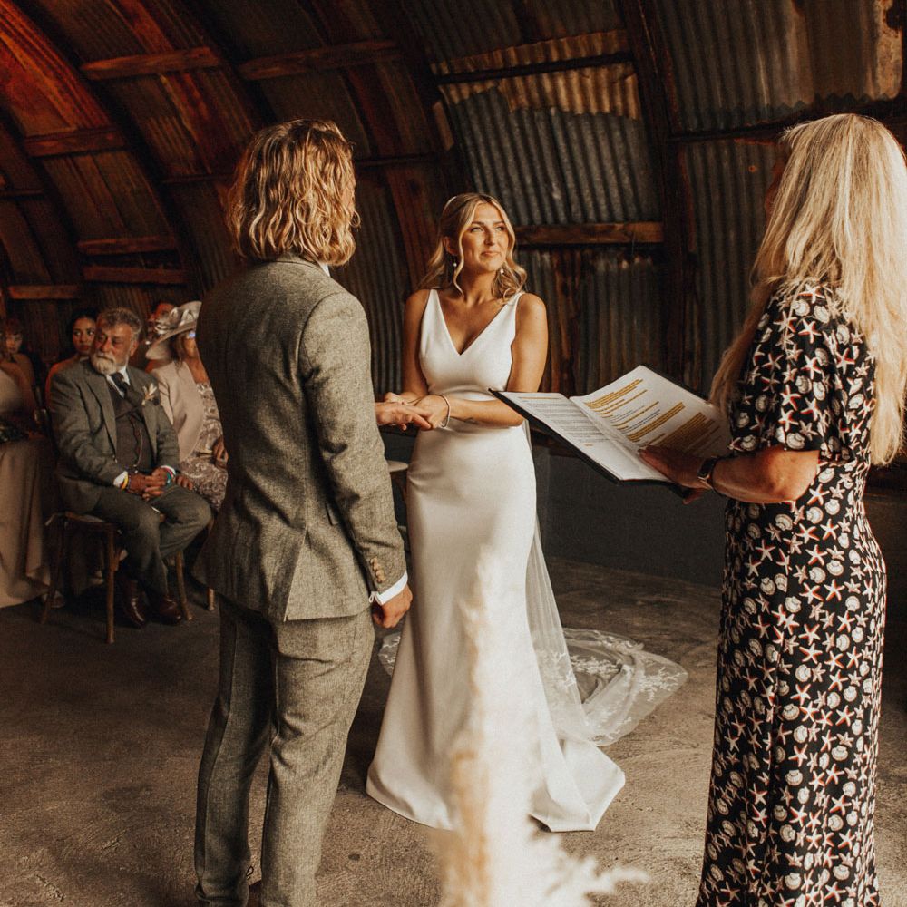 Bride in white V neck Made With Love wedding dress and veil stands holding hands with groom in sage green suit and brown shoes during bespoke wedding ceremony at Anran Devon