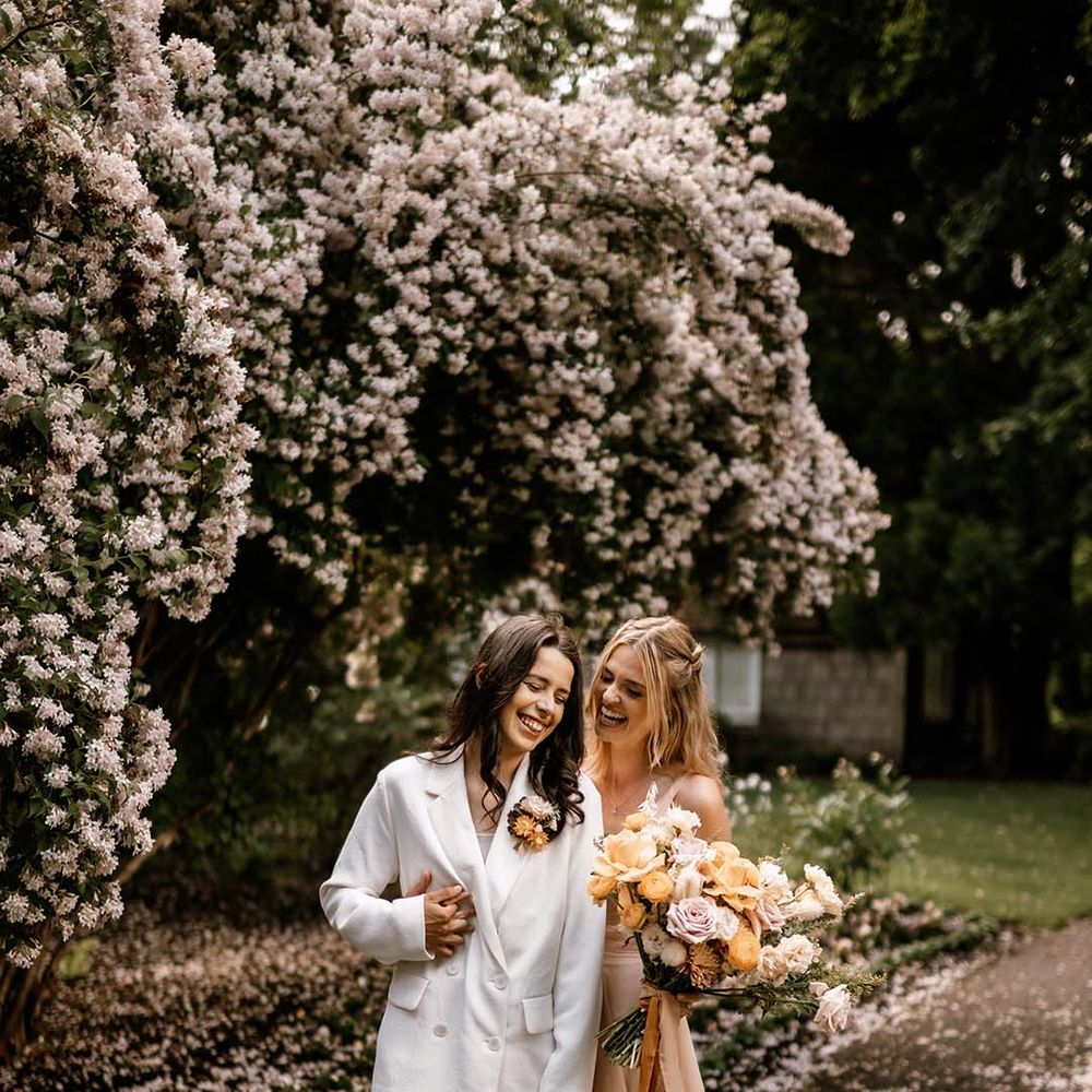 Bride in a blush pink tulle wedding dress holding a honey yellow and pink bouquet laughing with her bride in a white suit 