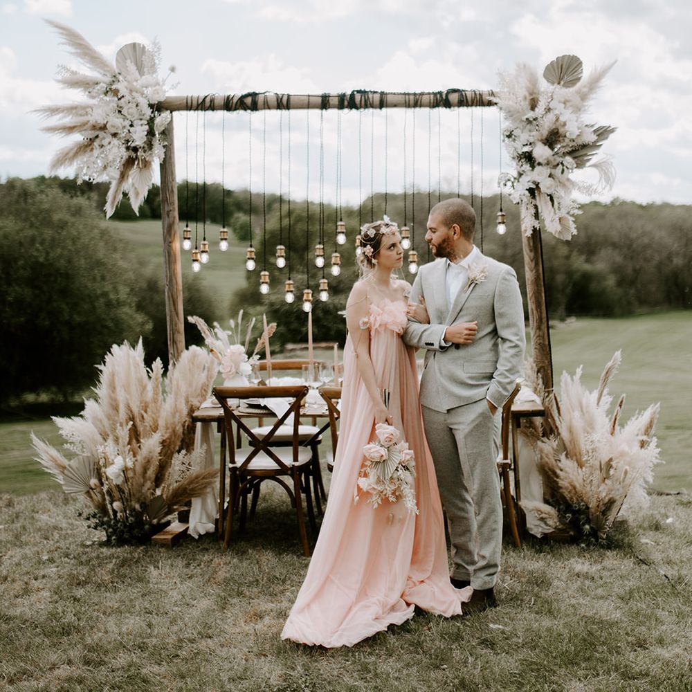 Boho luxe Waresley Park Estate wedding inspiration with bride in a blush pink wedding dress standing in front of a tablescape with pampas grass and Edison bulb installation
