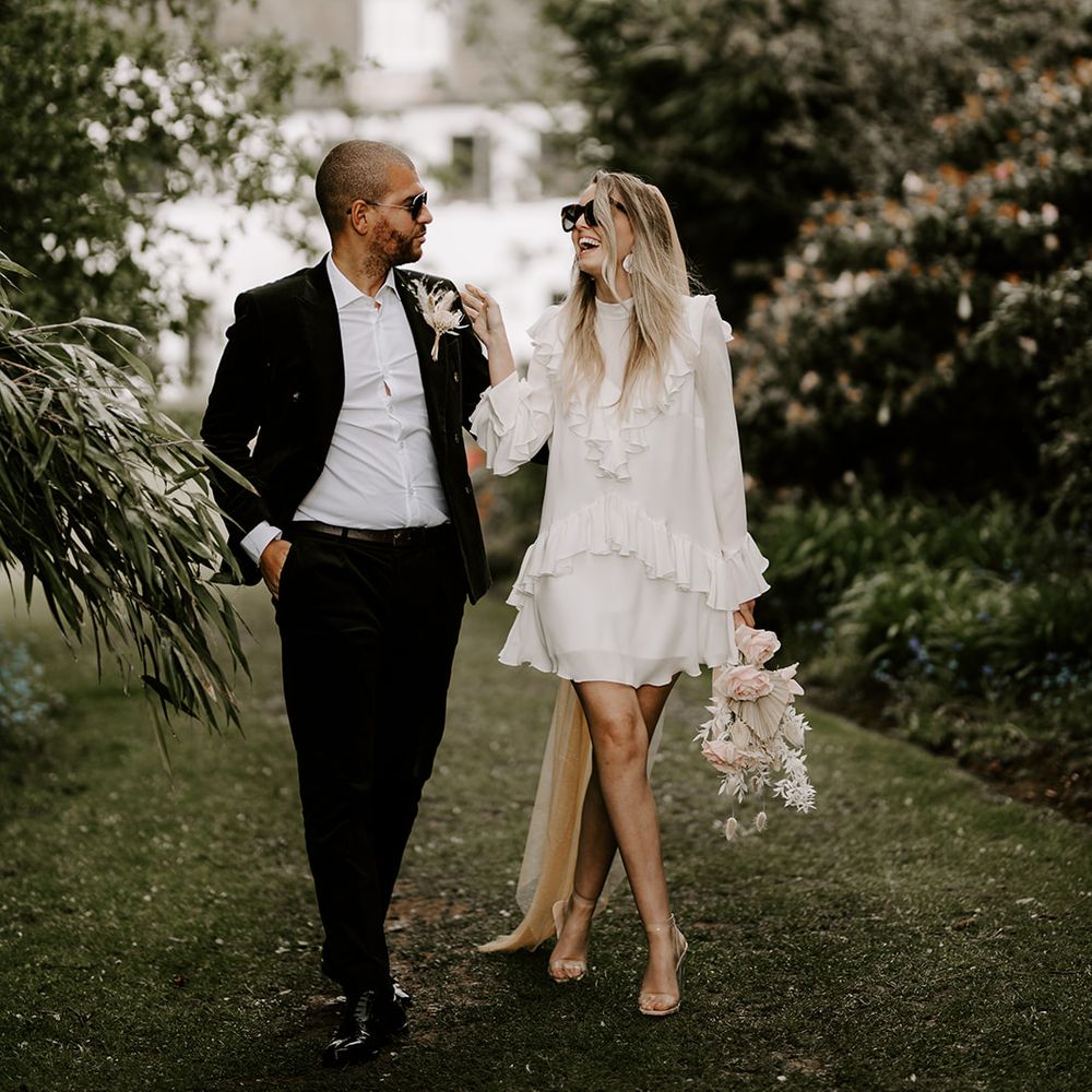Stylish bride in a short wedding dress, clear shoes and sunglass walking through the grounds of Waresley Park Estate with her groom in a black suit