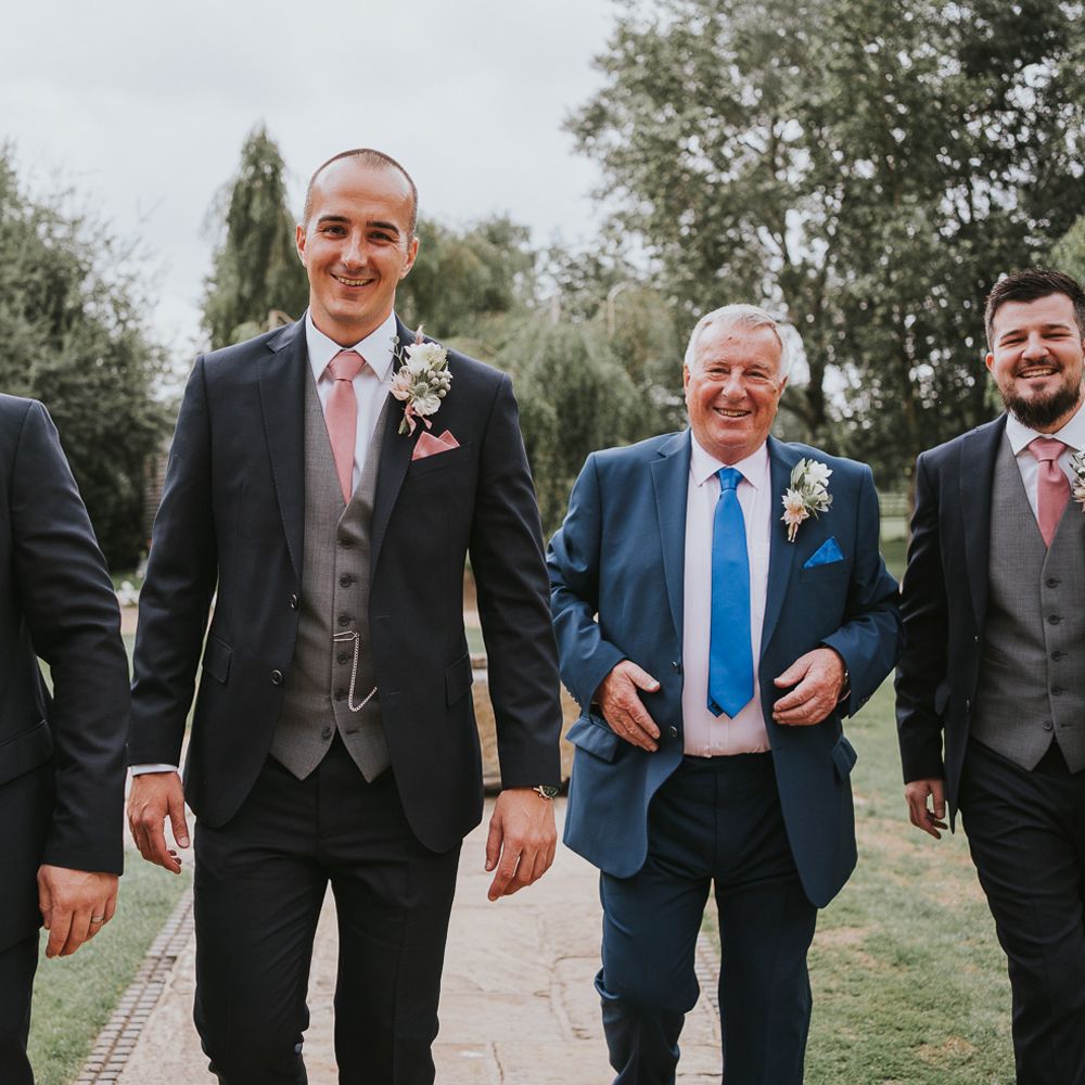 Groom and groomsmen in dark three piece suits with grey waistcoats, pink ties, pink pocket squares and protea buttonholes walking through gardens before Tythe Barn wedding with barn wedding flowers