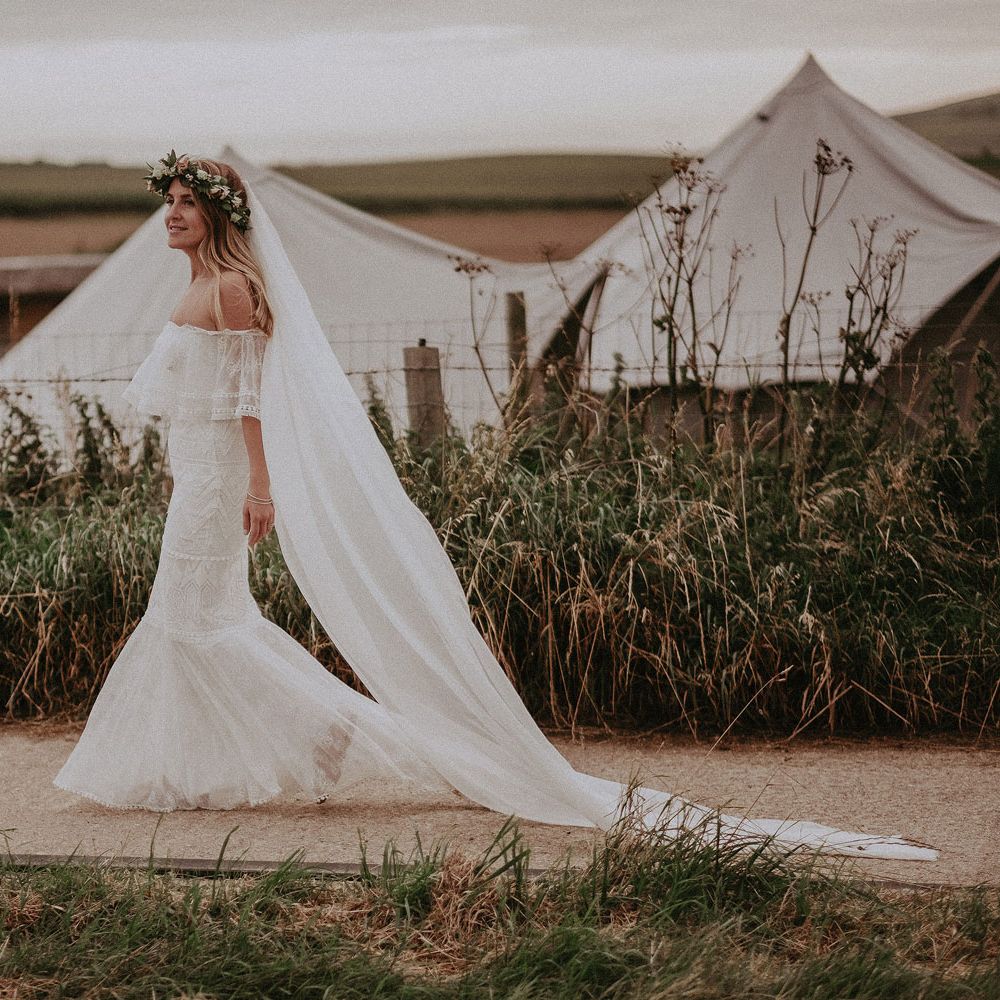 Bride in Grace Loves Lace bardot wedding dress, veil and flower crown walks along path by white tipis at Isle of Wight wedding with macrame wedding decor