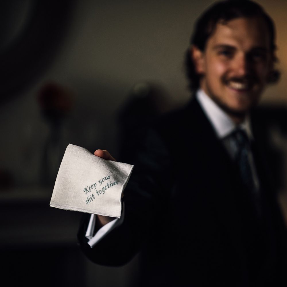Groom in three piece suit holds handkerchief with 'Keep your shit together' embroidered in green thread for Cornish wedding 