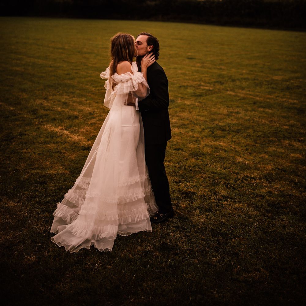 Bride in white Halfpenny London Mayfair dress with ruffle sleeves kisses groom in suit in field at garden wedding reception in Cornwall