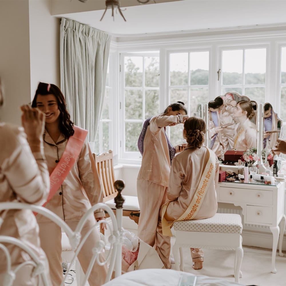 Bridesmaids in matching satin pyjamas and sashes have their make up done in bedroom before DIY garden wedding in Bedfordshire 