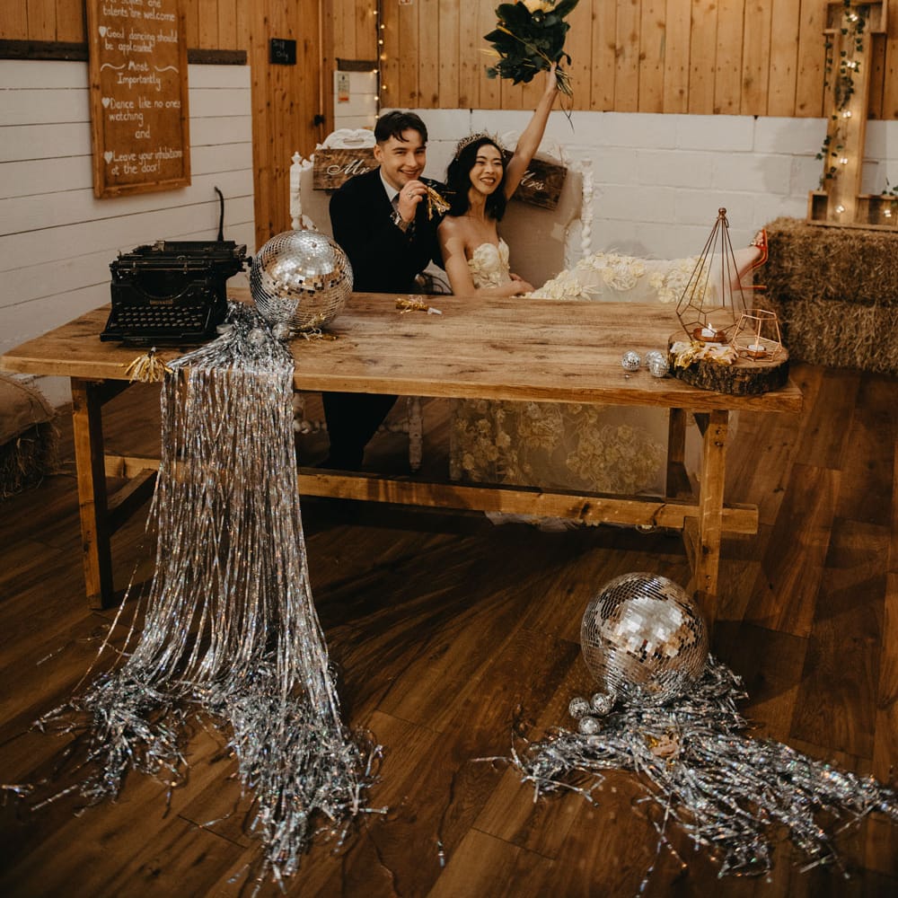 Bride in strapless feathered wedding dress and headpiece holds up bouquet as she sits with groom at wooden table for rustic wedding in Lancashire