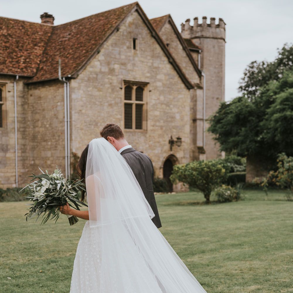 Bride in Pronovias wedding dress and cathedral veil walks through grounds of Notley Abbey holding white and green bridal bouquet with groom in grey suit