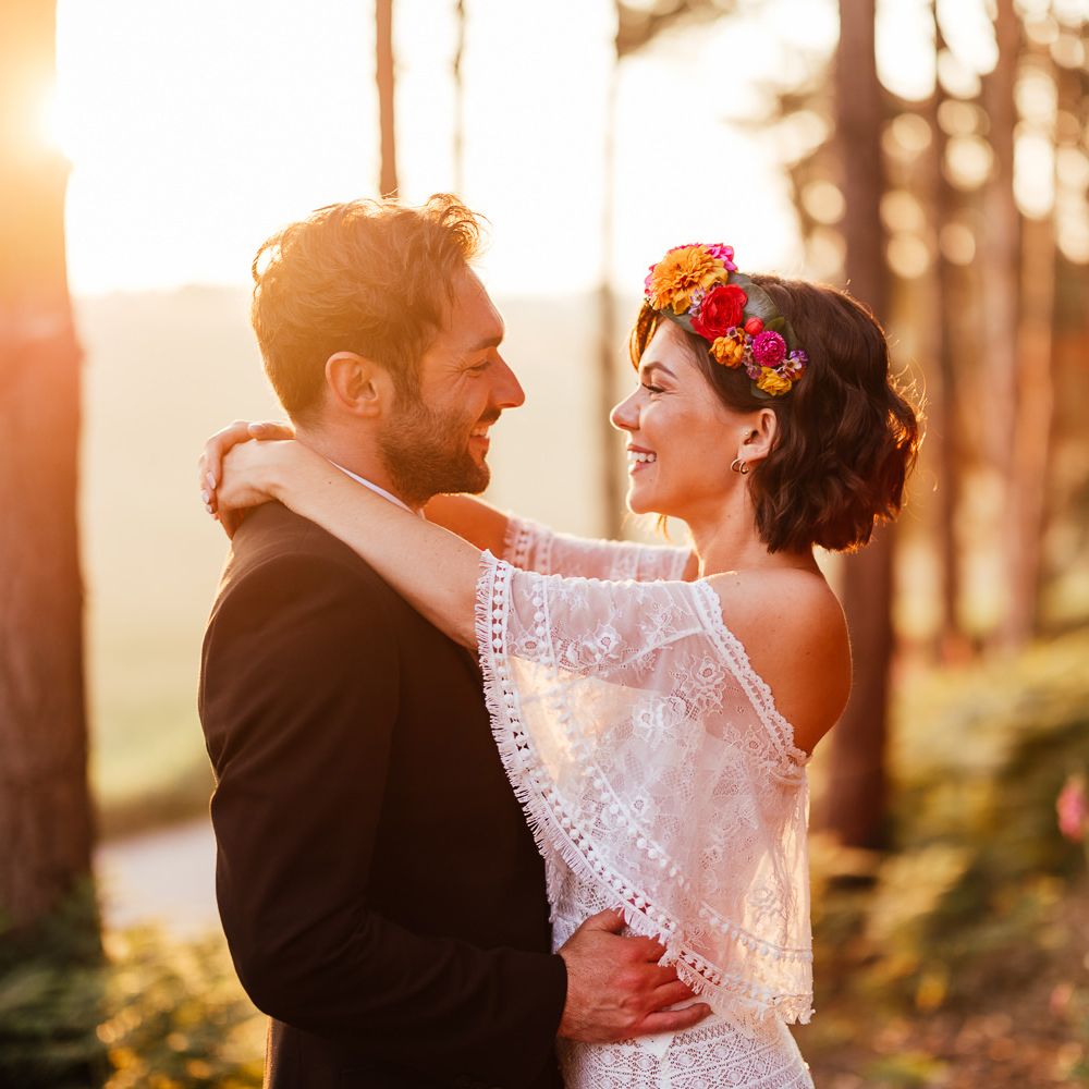 Smiling bride in off the shoulder Grace Loves Lace wedding Dres sand flower crown stands with smiling groom in black suit in woodland during golden hour