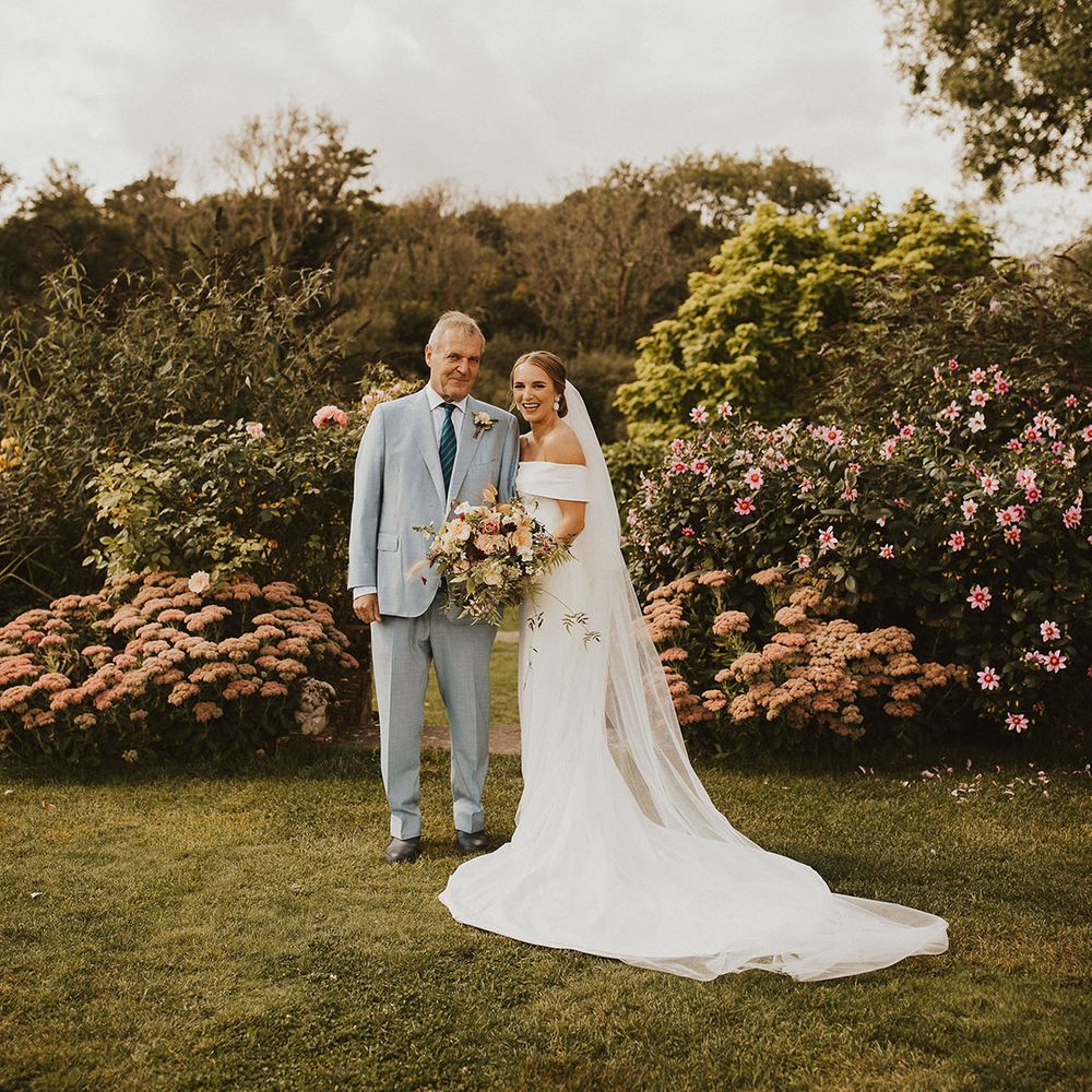 Bride stands with her father on her wedding day outdoors who wears pale blue suit