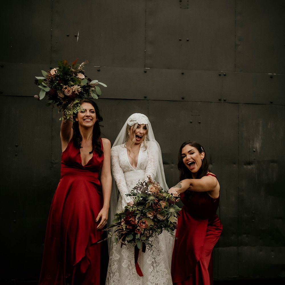 Boho bride in a lace wedding dress and Juliet cap veil standing with her bridesmaids in red satin bridesmaid dresses