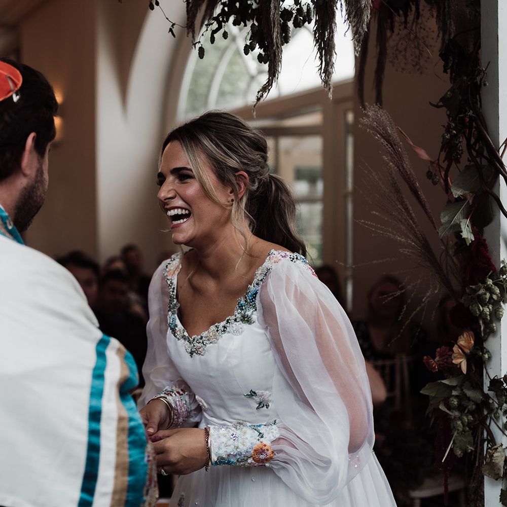 Laughing bride in mesh sleeve wedding dress with floral embroidery stands with groom in red kippah and tallit during wedding ceremony at Iscoyd Park