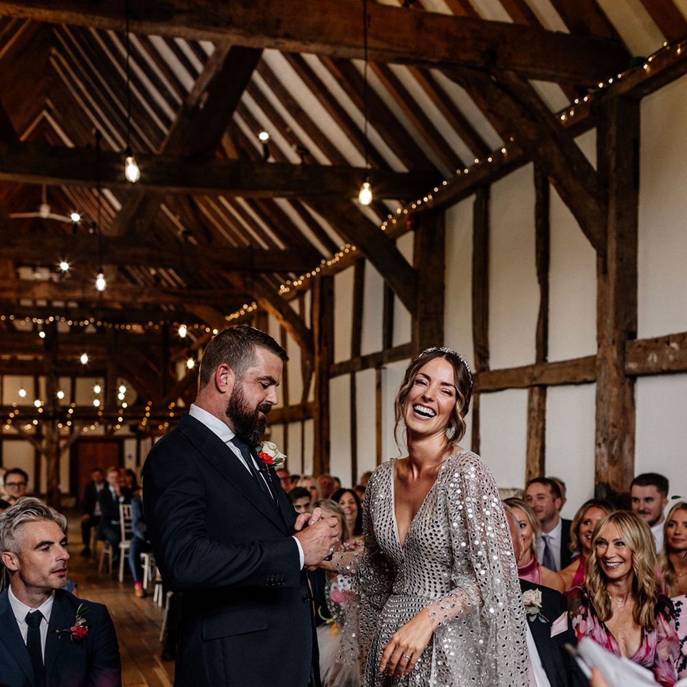 Groom in a navy suit holding hands with his bride in a silver sequin wedding dress at the altar of their rustic barn wedding 