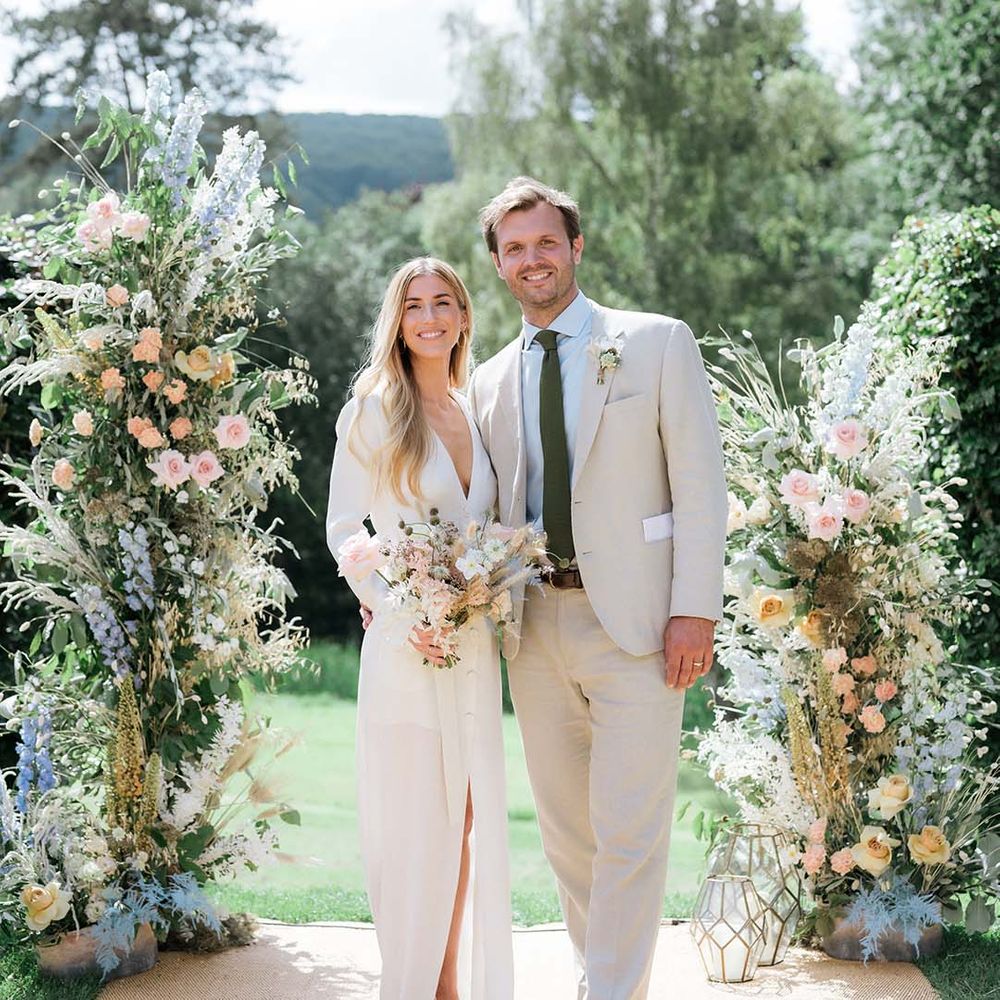 Bride in Bon Bride dress with plunging neckline with groom in cream suit in front of large pastel coloured flower displays