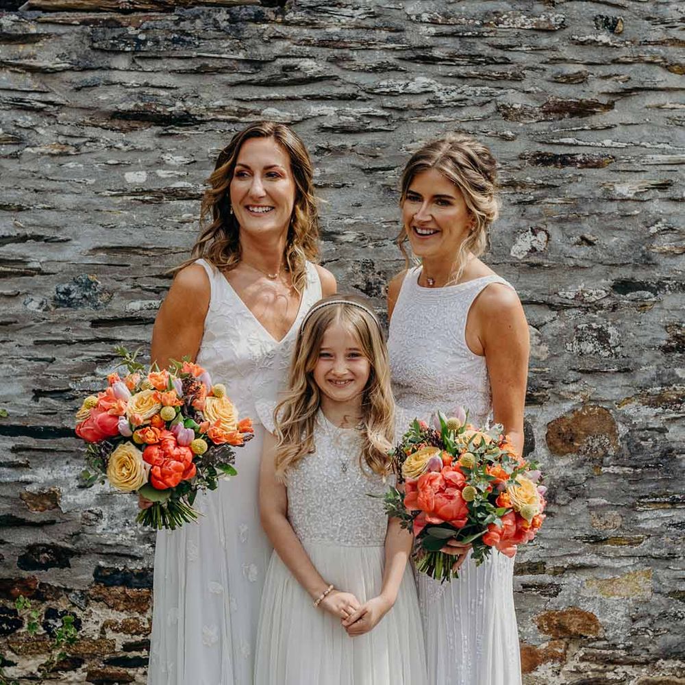 Two brides in tulle and lace embellished wedding dresses standing with their daughter holding colourful orange and yellow wedding bouquets 