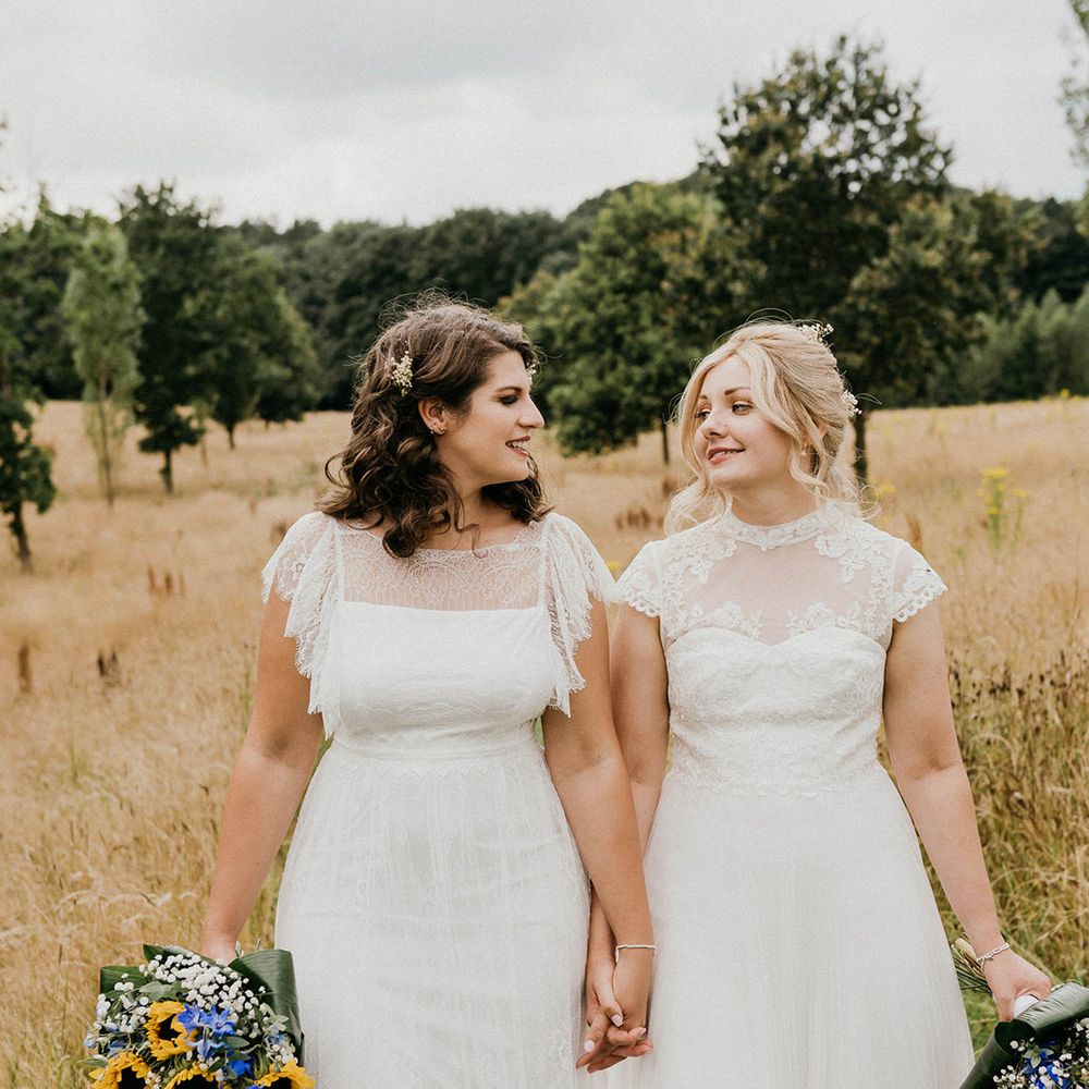 Brides glance at each other and hold hands as they walk with colourful bouquets