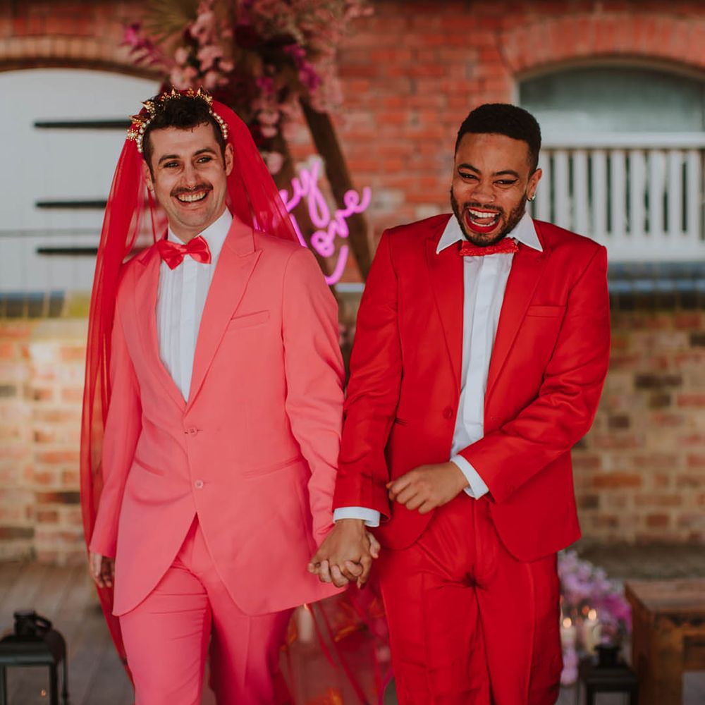Groom in a pink suit, bow tie, gold crown and red veil laughing and holding hands with his partner in a red suit and bow tie at Prestwold Hall Barns 