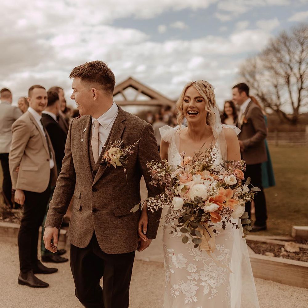 Bride in lace wedding dress with groom in brown three piece suit have confetti exit from barn wedding ceremony 