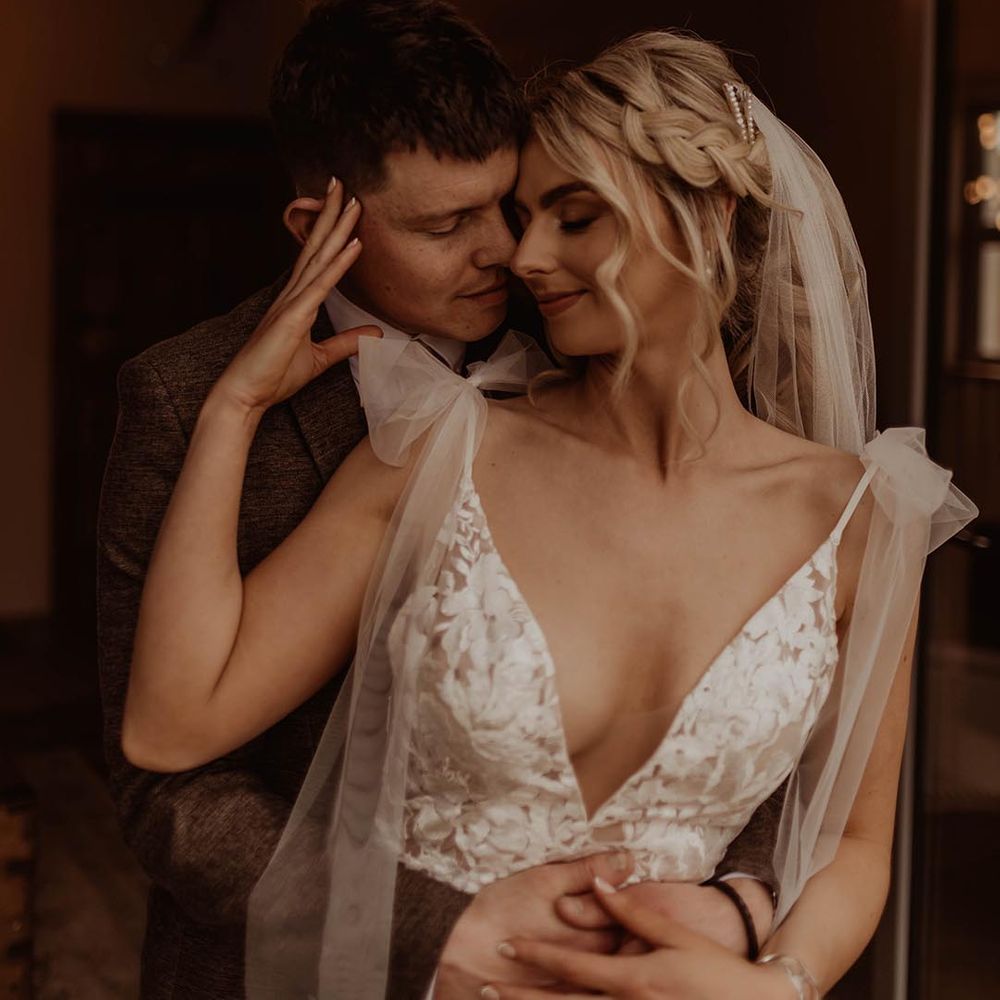 Bride in v neck lace wedding dress with bow sleeves and pearl earrings and diamanté hair clip with groom for rustic barn wedding 