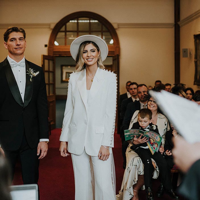 Bride and groom stand at the altar with groom in black tie with pale blue bow tie and bride in pearl trouser wedding suit 