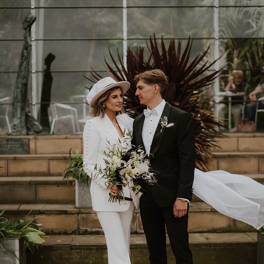 Bride and groom stand together with bride in trouser suit and groom in black tie 