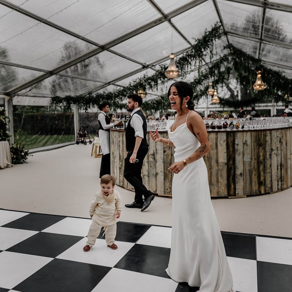 Bride in a satin slip wedding dress dancing on the dance floor of her clear glass marquee reception with her son 