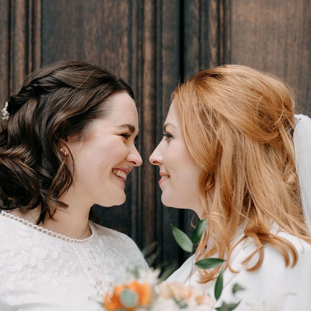 Brides with brown hair and red hair look lovingly at one another on their wedding day