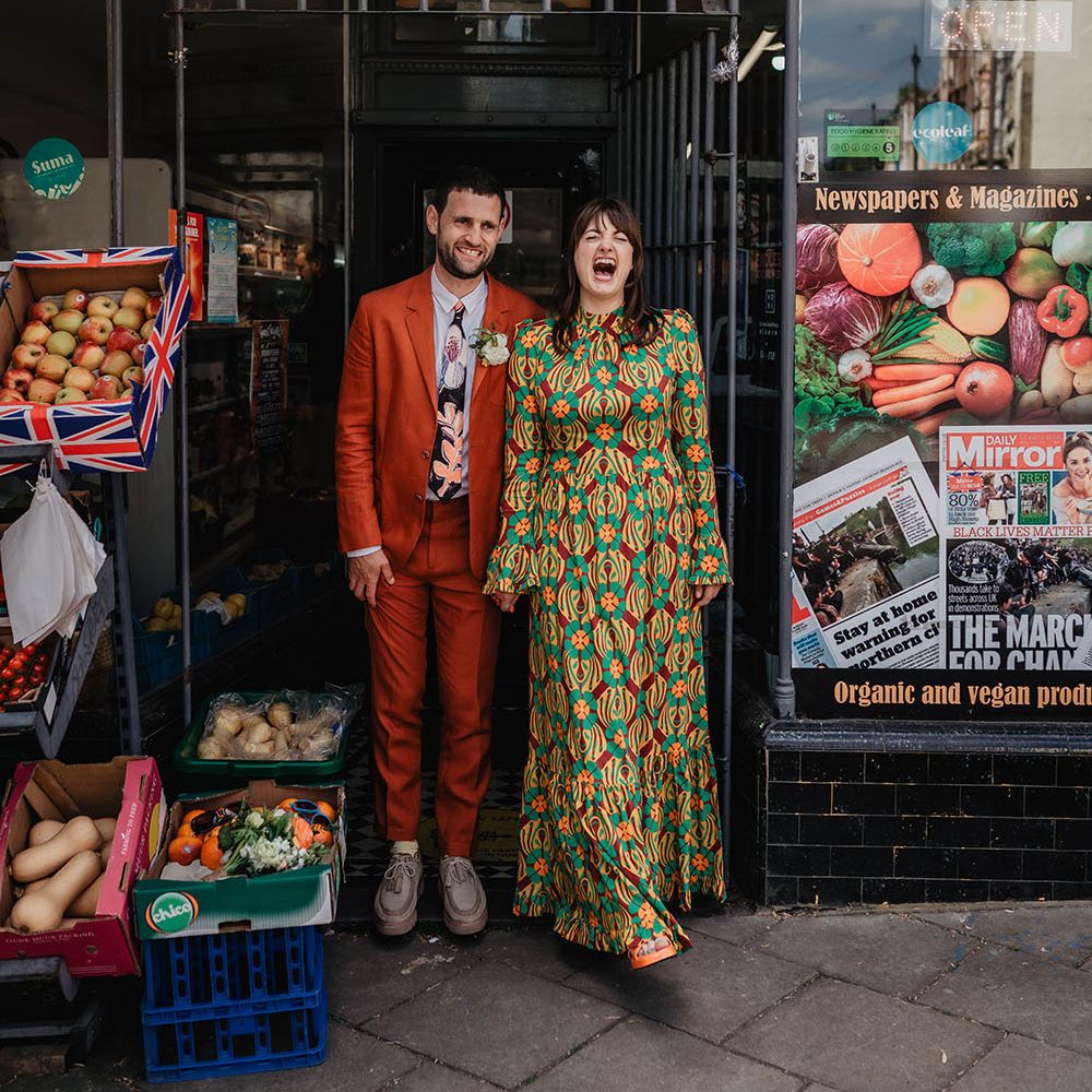 Bride & groom stand in shop doorway on their wedding day wearing brightly coloured outfits 