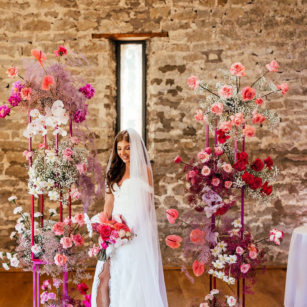Bride wearing off the shoulder lace wedding dress holds bright pink floral bouquet and stands in front of bright floral wedding arch 
