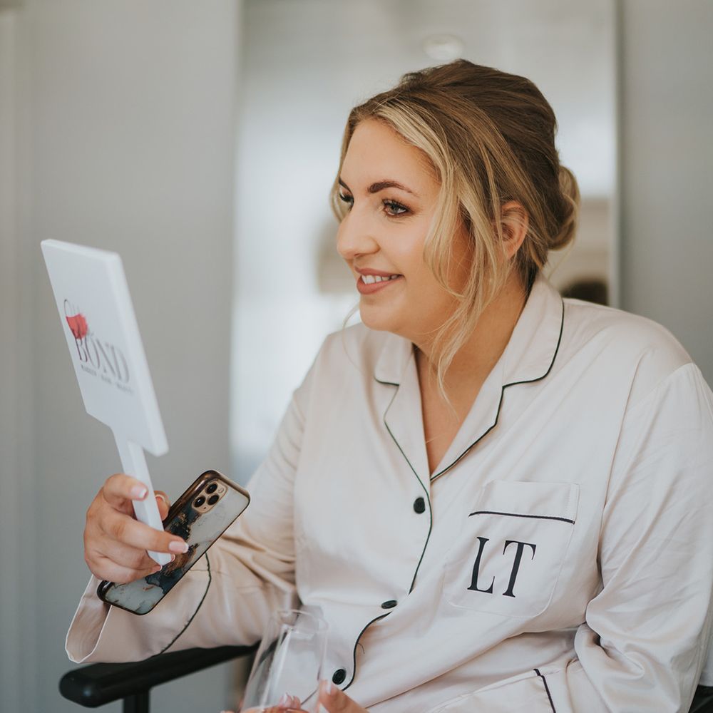 Bride in white pyjamas with personalised lettering looks at herself in the mirror after having her wedding makeup done