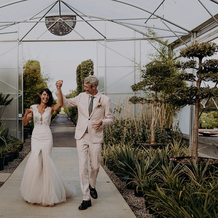 Bride and Groom walk through greenhouse holding hands in the air and smiling