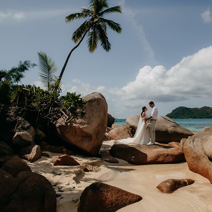 Bride in off the shoulder lace wedding dress with sweetheart neckline and groom in crisp white shirt, light coloured chinos, tan leather belt and white trainers embracing on large rock on a beach in Seychelles with palm trees and azure coloured sea