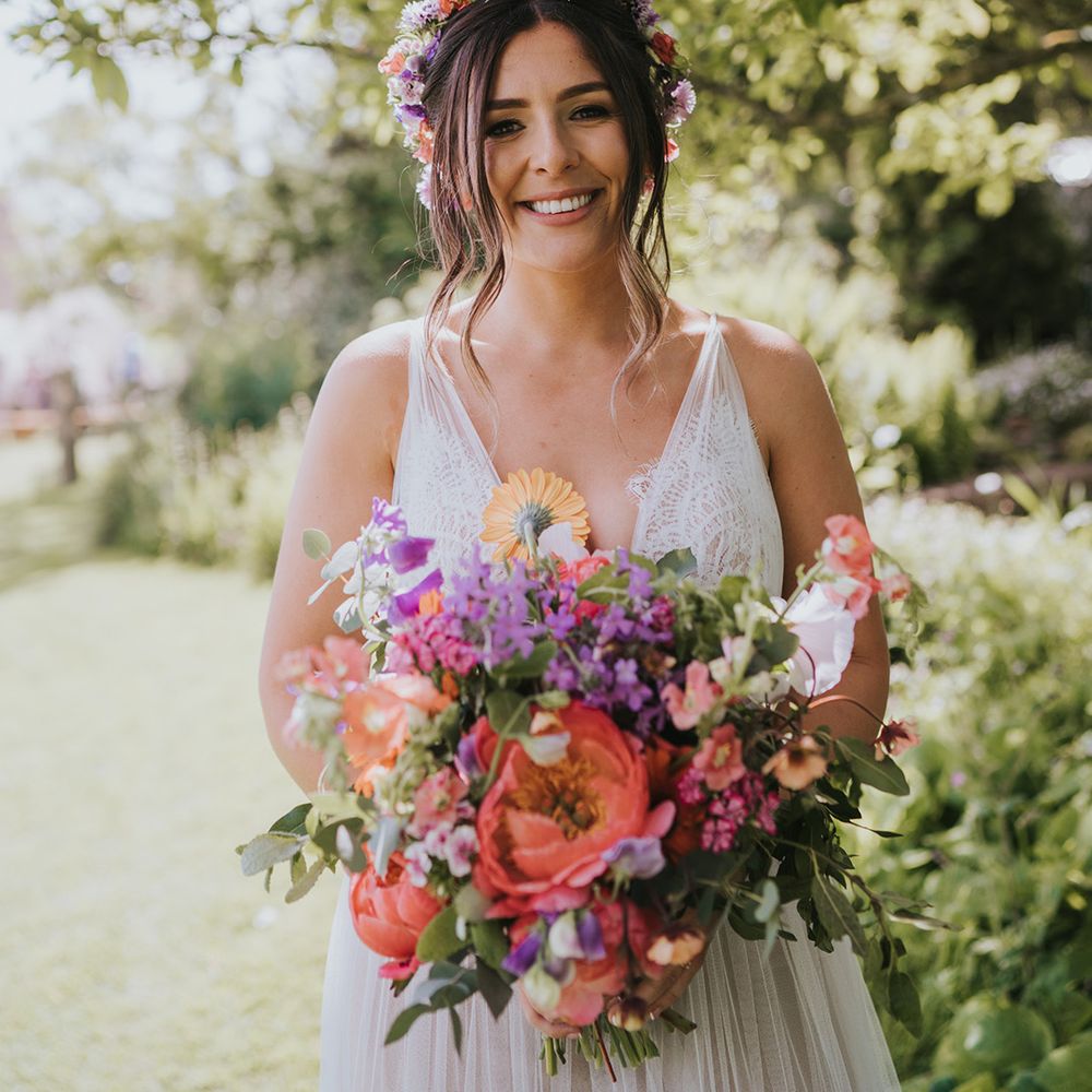 Bride wears scalloped lace wedding dress and colourful flower crown whilst holding matching bouquet 