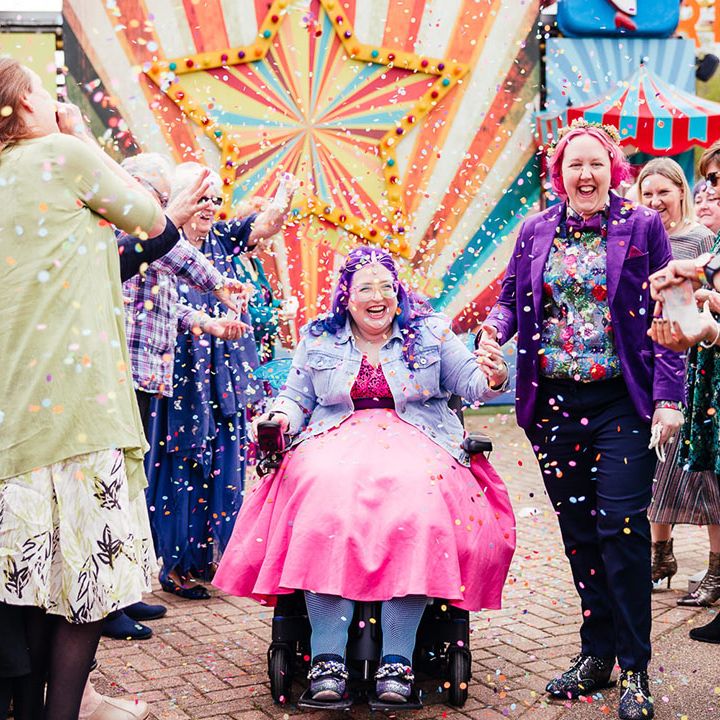 Bride in purple velvet blazer and black suit trousers with purple bowtie, pink patterned pocket square and colourful patterned shirtband bride in wheelchair wearing personalised denim jacket, pink wedding dress and blue tights doing confetti walk at theme park wedding 