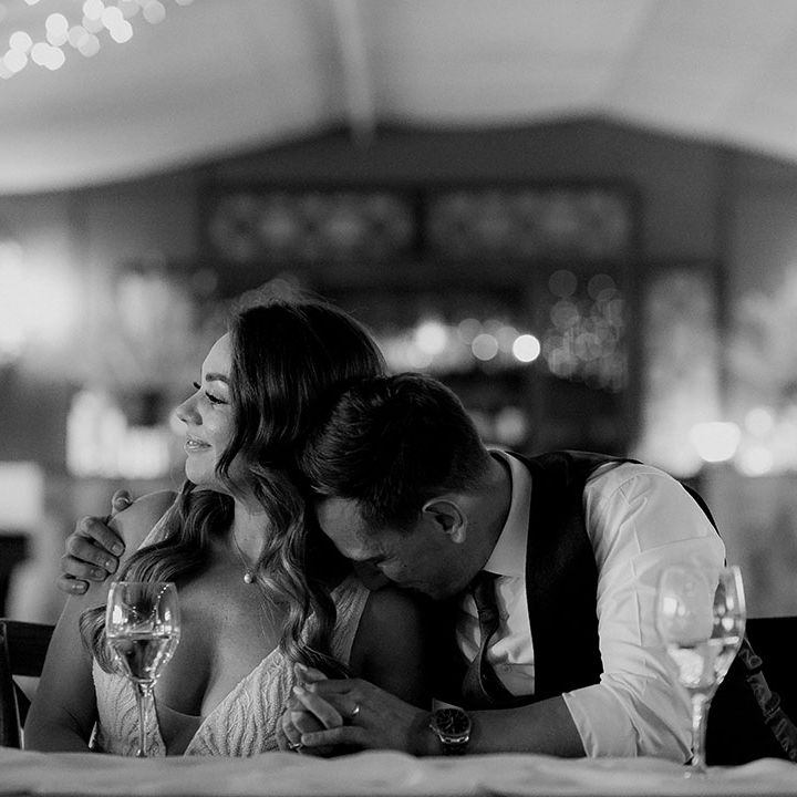 The groom kisses the bride on her shoulder as they sit at their wedding reception as they listen to speeches 