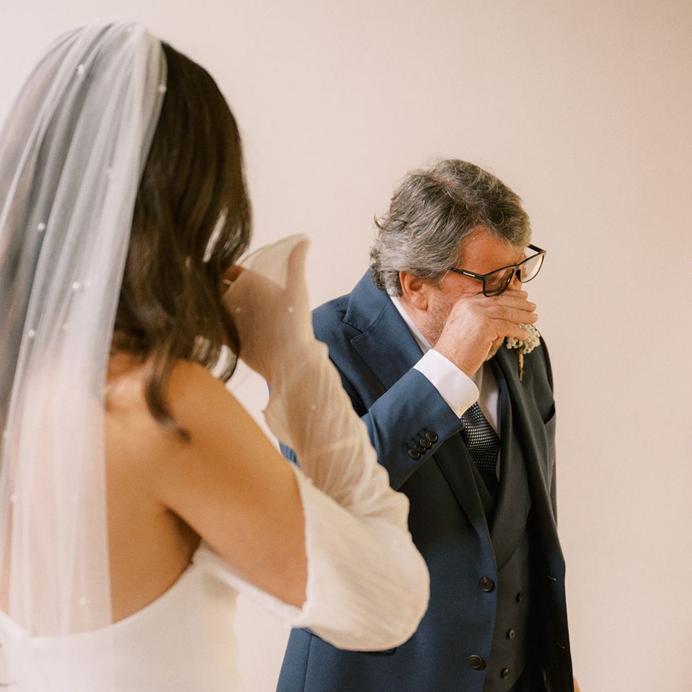 Father of the bride in a three piece navy suit wiping his eyes as he cries seeing the bride in a pearl embellished bridal outfit 