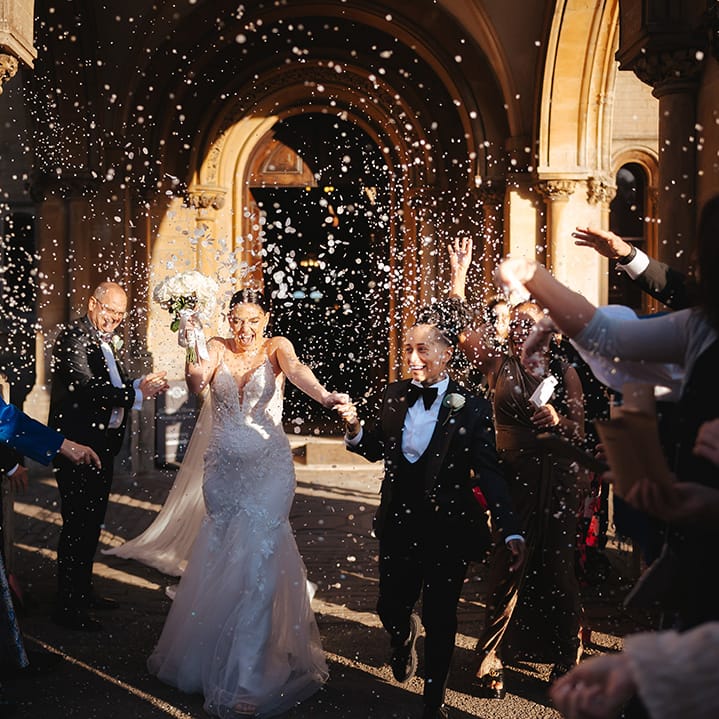 Bride in black snakeskin print tuxedo with black bowtie and white rose boutonniere holding hands with bride in lace sleeveless v neck Enzoani wedding dress with tulle finish and church length veil holding white garden rose bridal bouquet doing white petal confetti exit at wedding