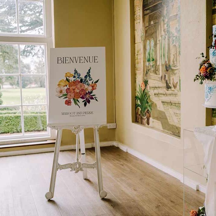Large botanical luxe white wedding sign in French on white easel next to four tiered blue and white wedding cake 