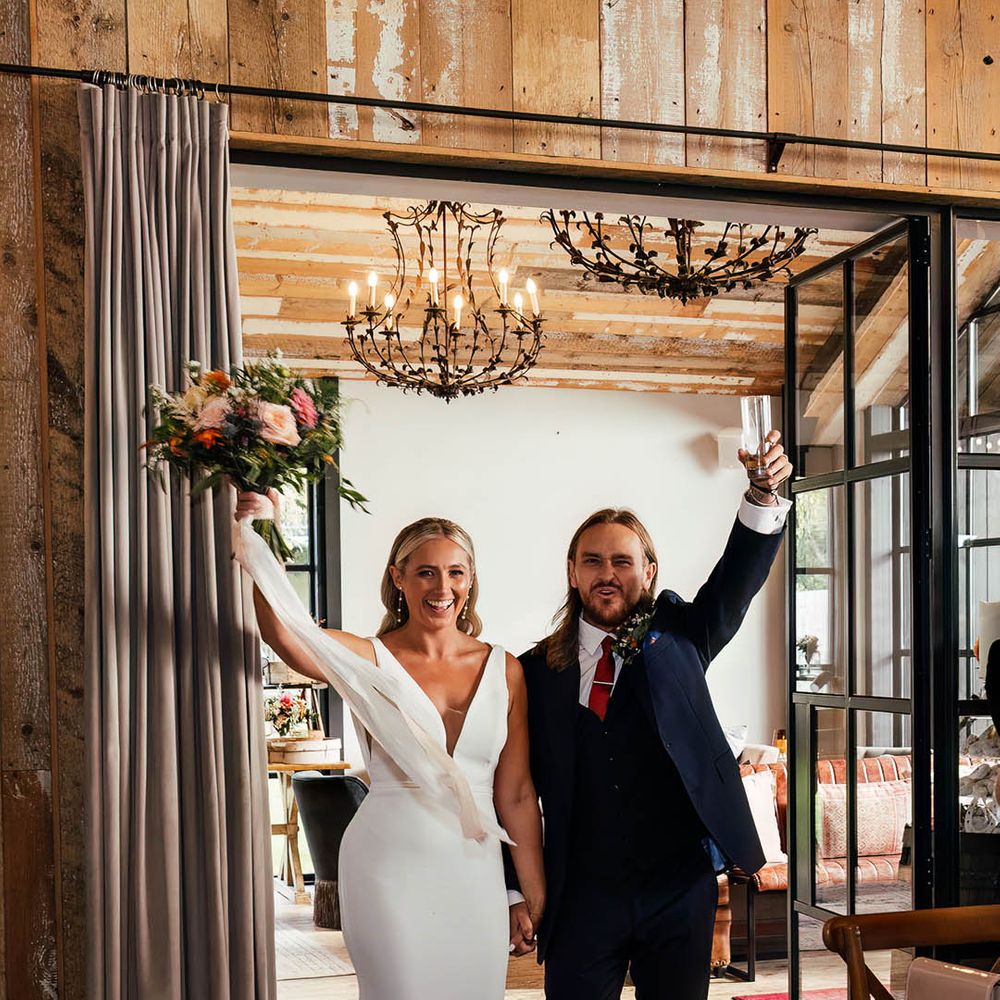 The bride in a fitted Savin London wedding dress enters the wedding reception with the groom in a three piece blue suit 