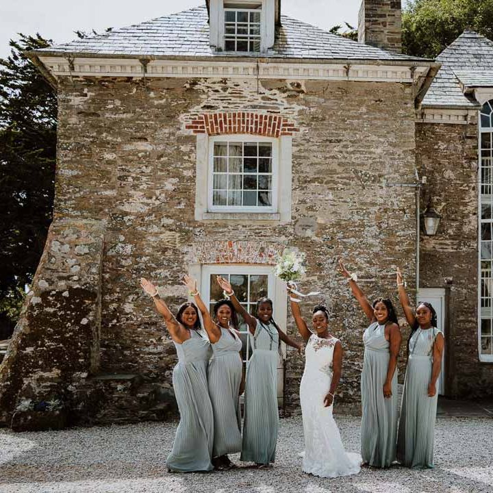 Bridesmaids in mismatched baby blue bridesmaid dresses standing with bride in lace and mesh boatneck wedding dress with silver bridal hair accessories holding up large white rose and foliage bridal bouquet outside of Tredudwell Manor wedding venue Cornwall