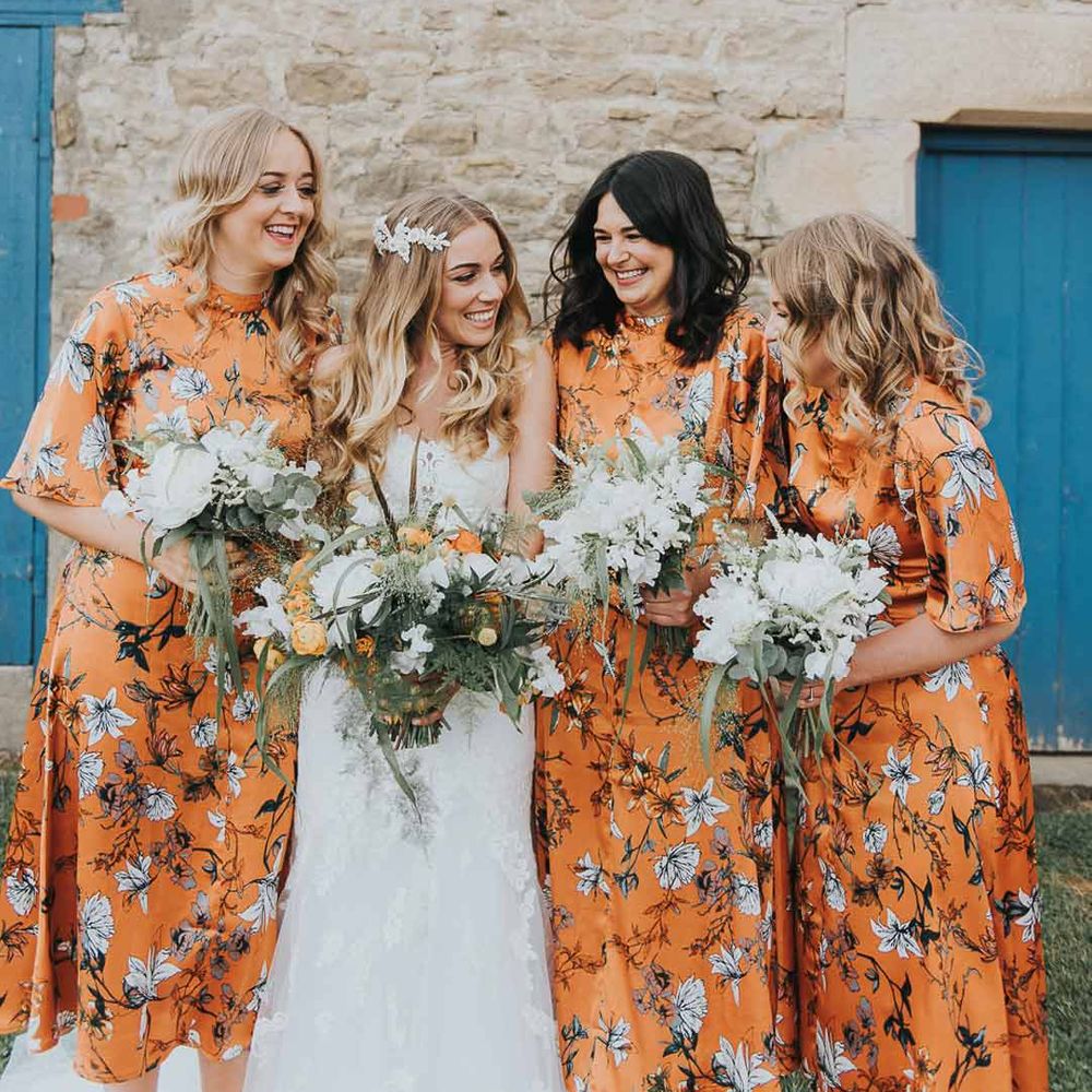 Bride in strapless lace wedding dress standing with bridesmaids in short sleeve midi orange floral patterned bridesmaid dresses