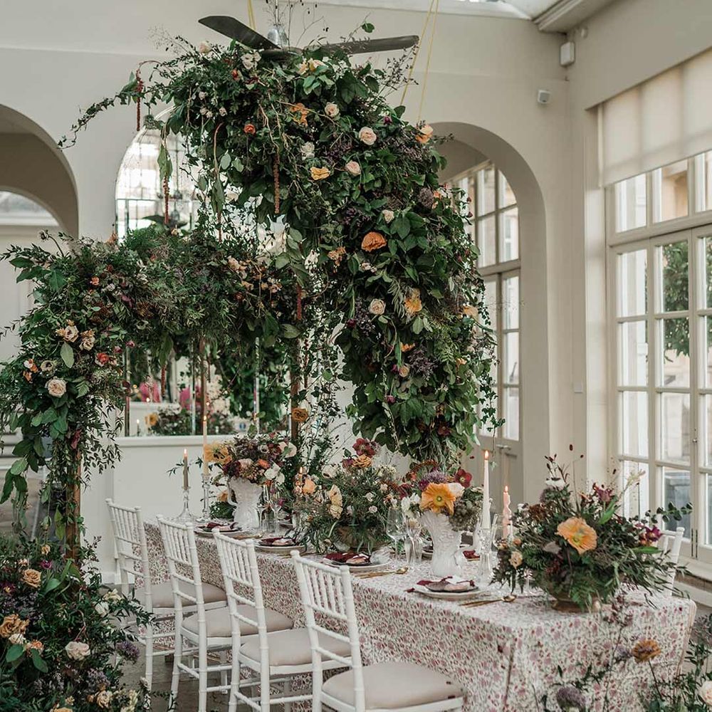 Botanical wedding tablescape with patterned floral tablecloth, twisted tapered candles, art deco vases with dried wildflower wedding centrepieces surrounded by suspended wildflower and foliage arrangements in large gold vases at Buxted Park