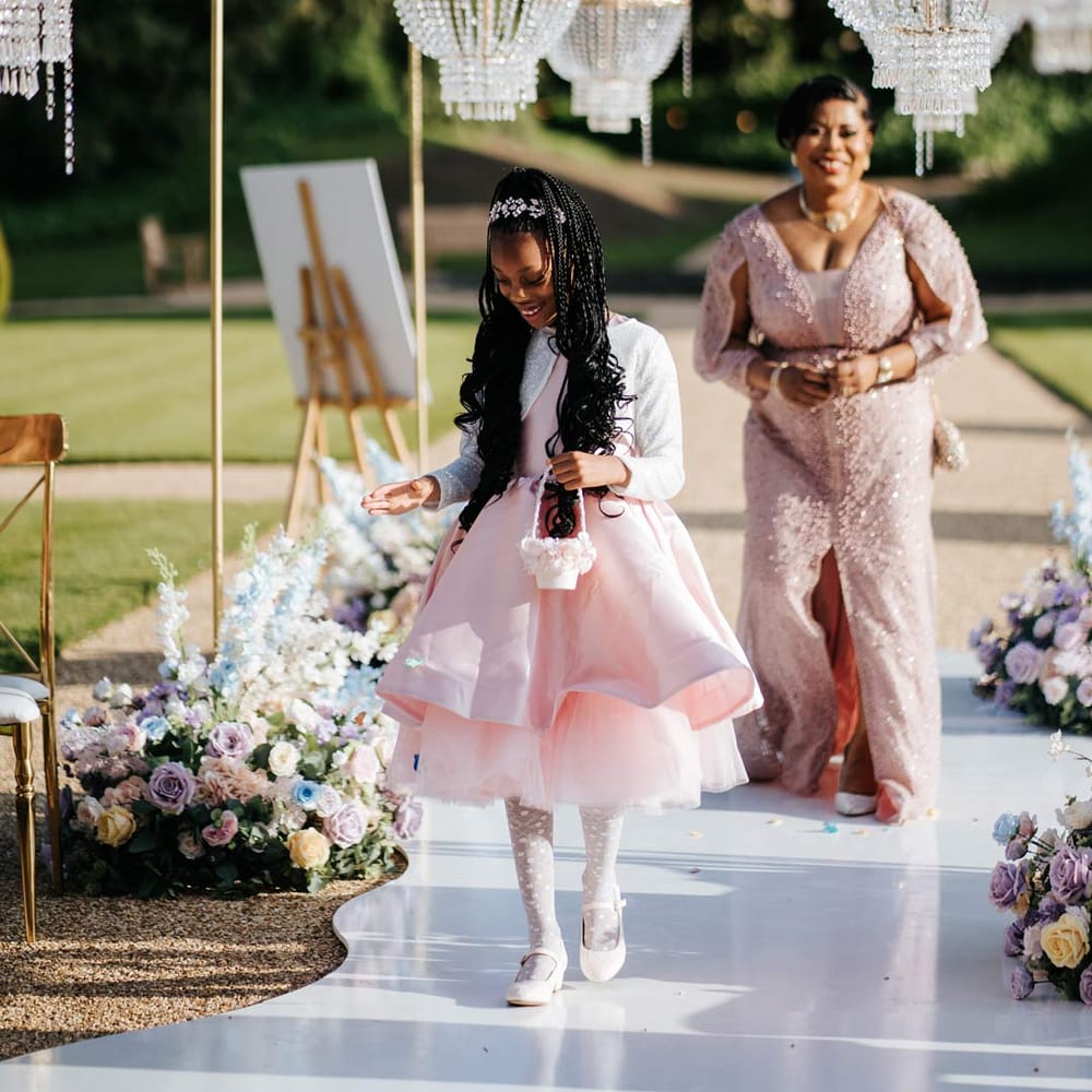 Flower girl in baby pink flower girl dress and white cardigan walking down the aisle at Syon Park wedding venue with outdoor gold and crystal chandeliers and pastel aisle flower arrangements with bride wearing illusion v neck sheer off shoulder long sleeve pearl embellished wedding gown walking behind her