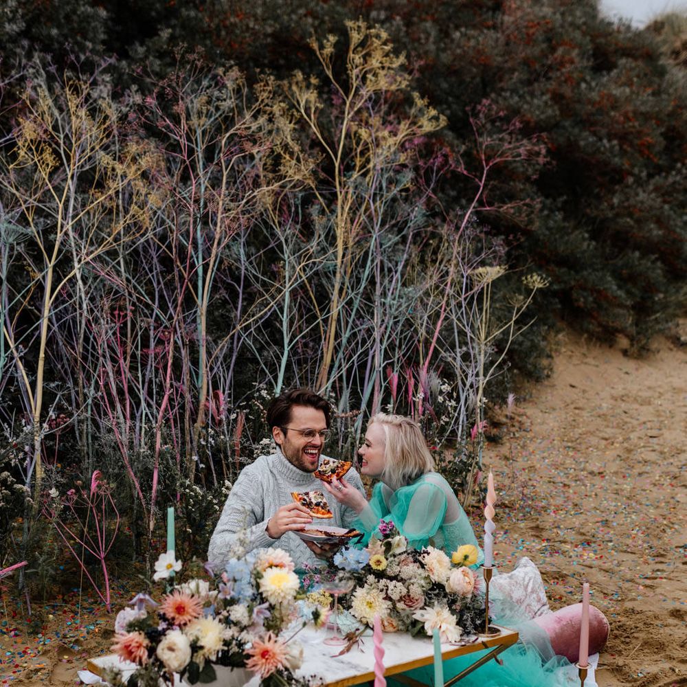 Intimate pizza picnic on the beach with pastel flowers and candles staked in the sand 