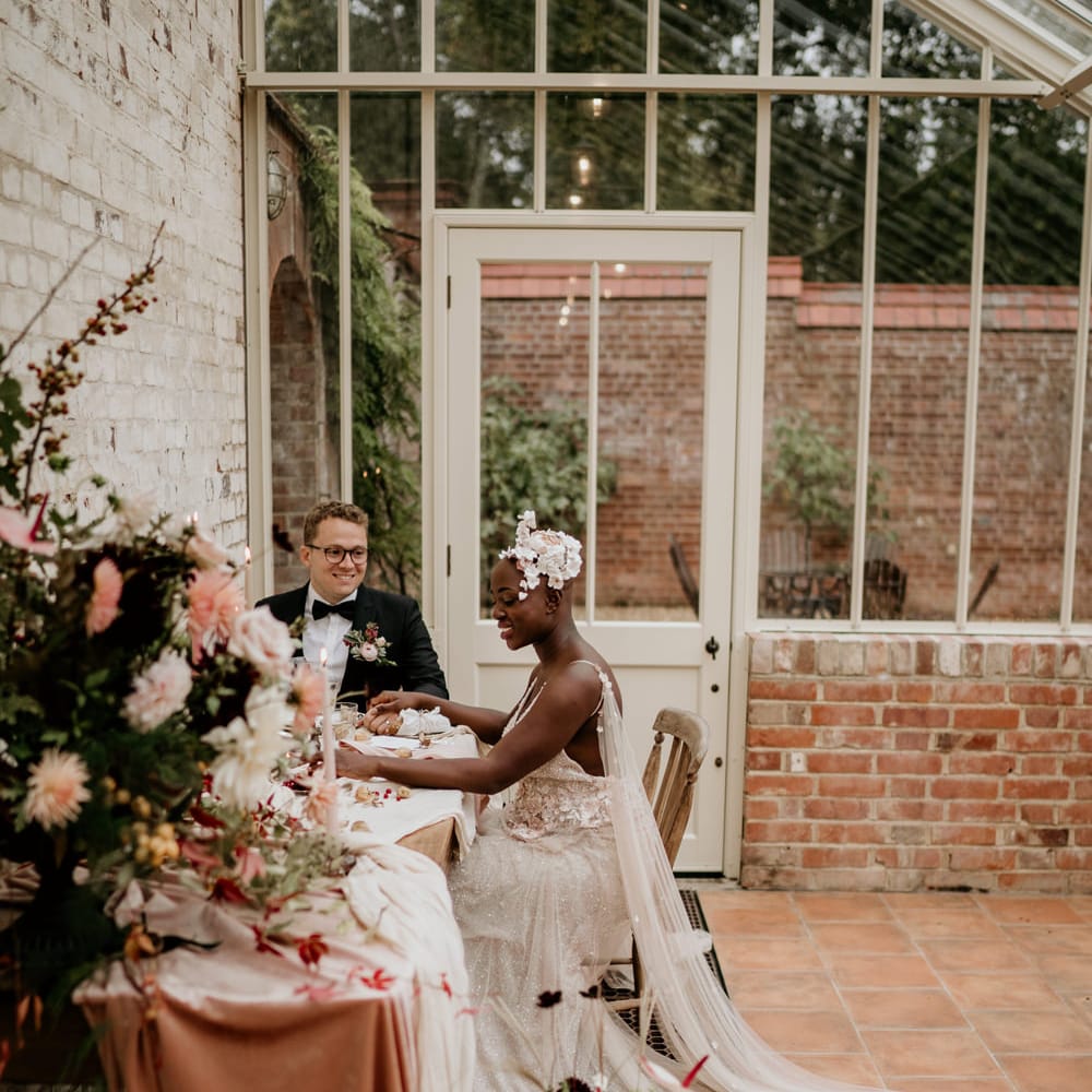 Bride with alopecia sitting at her intimate wedding breakfast in Our Beautiful Glasshouse in Sussex in a blush Julita Bridal London with watteau train 