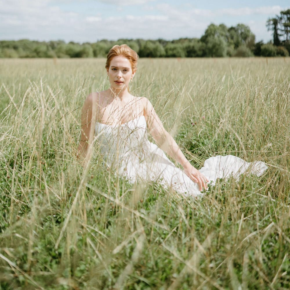 Bride sitting in the fields at Berwick Lodge wearing a spaghetti strap wedding dress, relaxed low pony tail and gold earrings