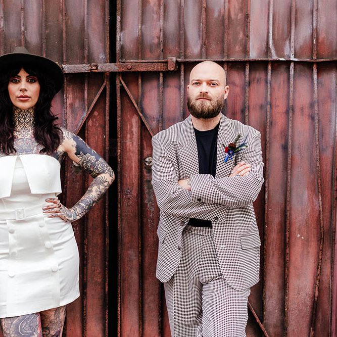 Bride in an alternative wedding dress with buttons wearing a fedora hat and standing next to a container with her groom in a check suit 