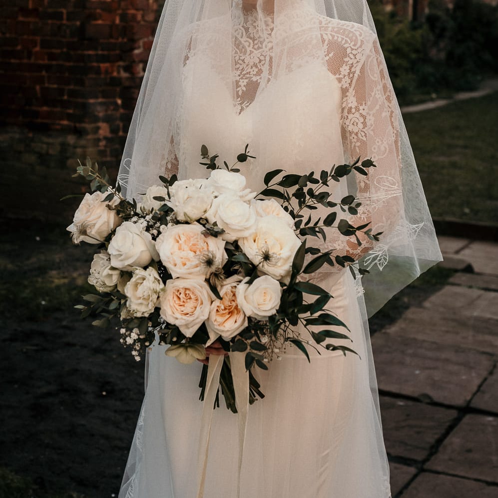 Classic bride in a long sleeve lace wedding dress with sheer veil holding an all rose wedding bouquet 
