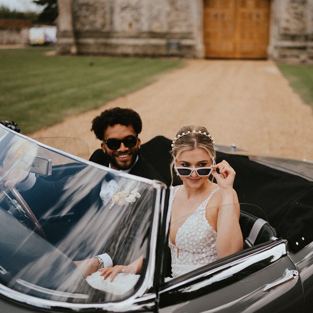 Bride in white sunglasses with a pearl headband sitting in a classic wedding car with her husband in a tuxedo 