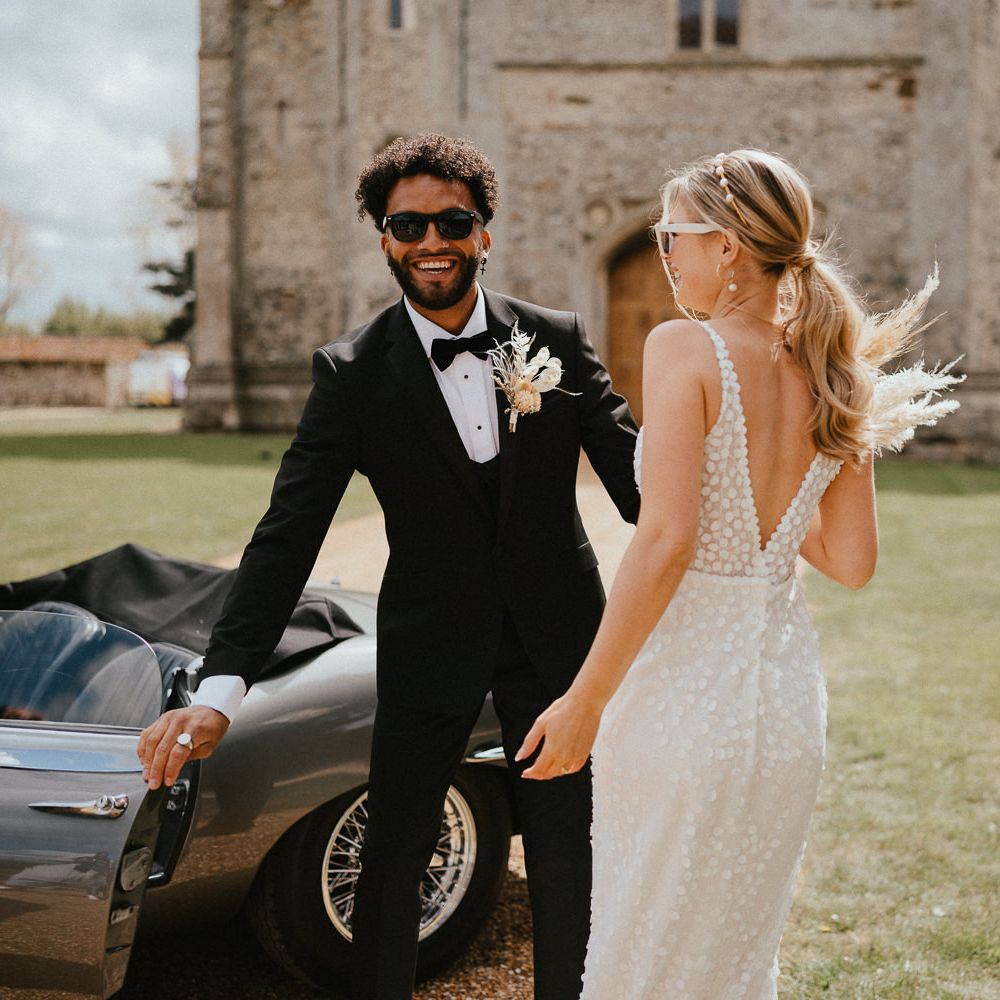 Groom in a tuxedo with bow tie, sunglasses and dried flower buttonhole holding the car door open for his bride in an appliqué wedding dress